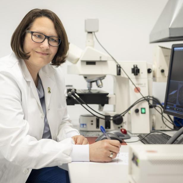 Person in weißem Laborkittel sitzt an Schreibtisch mit Mikroskop und Computerbildschirm mit wissenschaftlichen Grafiken