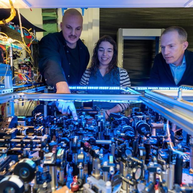 Drei Personen stehen an einem komplexen optischen Experimentaufbau mit zahlreichen Linsen, Spiegeln und Kabeln in einem Labor.