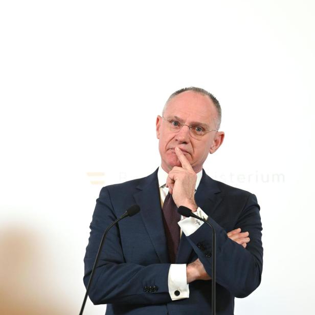 Ein Mann im Anzug (Innenminister Karner) steht nachdenklich mit verschränkten Armen und Finger am Mund vor zwei Mikrofonen.