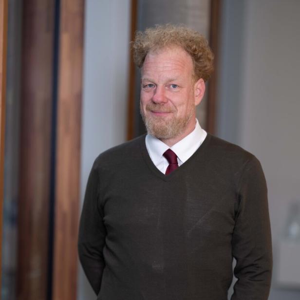 Ein Mann mit lockigem, blondem Haar und Bart trägt einen dunkelgrünen Pullover über einem Hemd mit Krawatte.