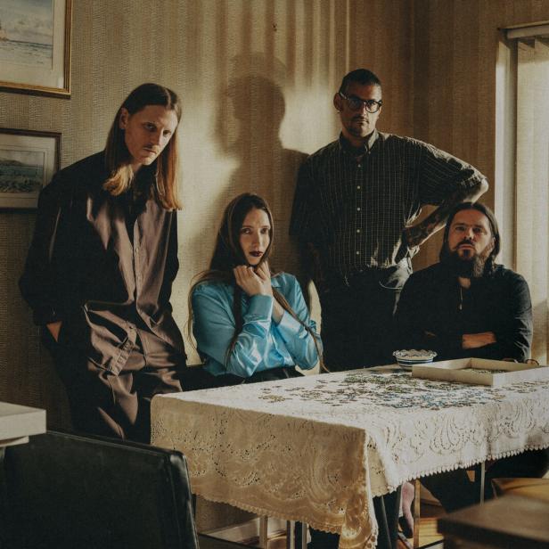Die Band Dry Cleaning, ernst an einem Tisch in einer altmodisch möblierten Wohnung sitzend und stehend