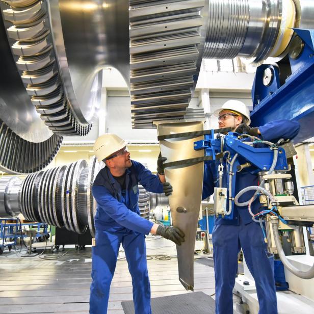 Zwei Arbeiter in blauer Arbeitskleidung und Schutzhelmen montieren ein großes Metallteil in einer Industriehalle mit großen Zahnrädern im Hintergrund