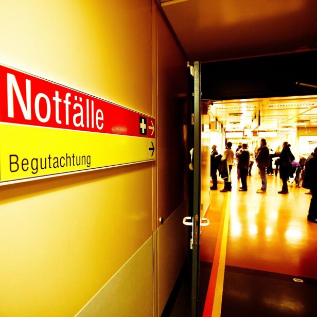 Ein Flur mit einem Notfall-Schild und mehreren wartenden Menschen in einem hell beleuchteten Bereich.