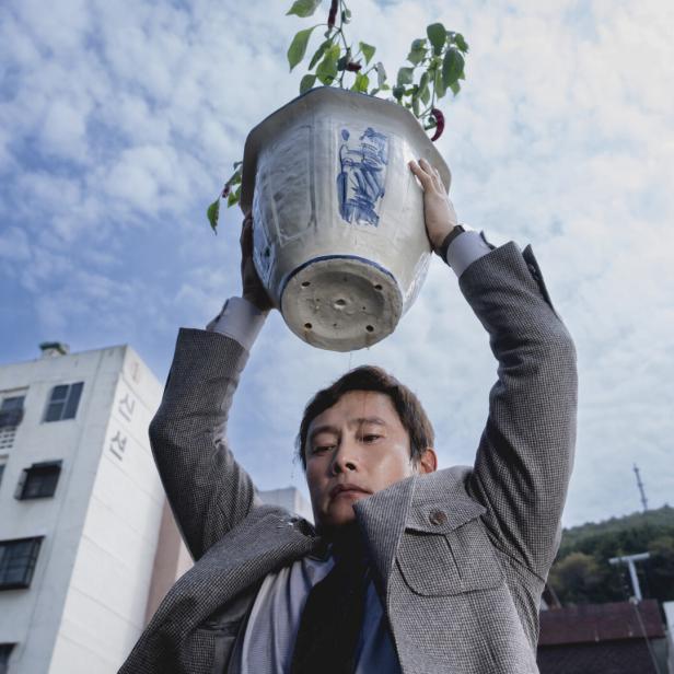 Der Schauspieler Lee Byung-hun, von unten gesehen, mit einem großen Blumentopf, den er wie wurfbereit über seinen Kopf hält 