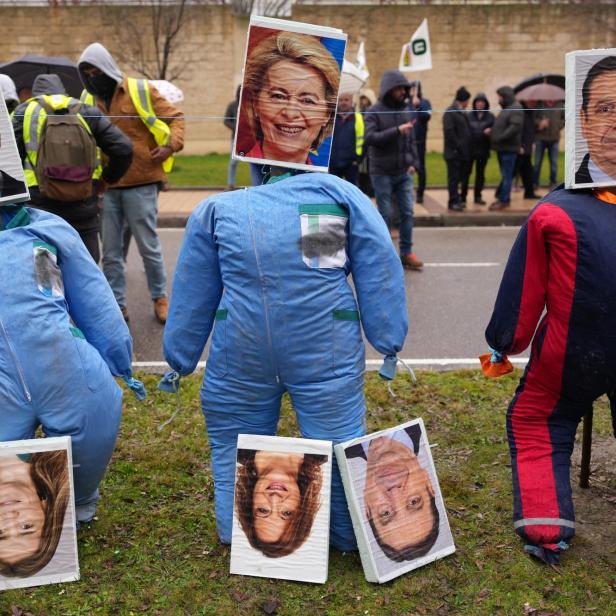 Mercosur-Protestierende stehen in Warnwesten und mit Schildern auf einer Straße. Im Vordergrund sind drei ausgestopfte blaue Ganzkörper-Anzüge zu sehen. Dort wo der Kopf rausschauen sollte, hängen stattdessen Bilder der am Abkommen beteiligten Staatsoberhäupter. Links Santiago Peña, Mitte Ursula von der Leyen, rechts Brasiliens Ex-Präsident Jair Bolsonaro