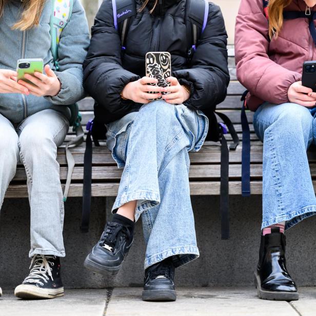 drei Mädchen sitzen auf einer Bank nebeneinander und schauen aufs Handy, ihre Köpfe sind abgeschnitten