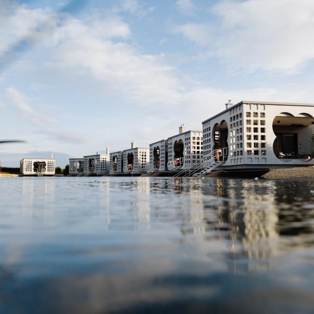 Reihe moderner, kubisch gestalteter Gebäude mit großen Öffnungen am Ufer eines ruhigen Gewässers unter blauem Himmel