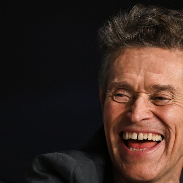 Der Schauspieler Willem Dafoe, hellauf lachend vor dunklem Hintergrund 