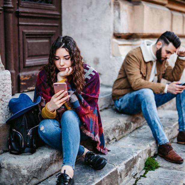 Eine Frau und ein Mann sitzen, voneinander abgewandt, auf Steinstufen, und starren auf ihre Smartphones.