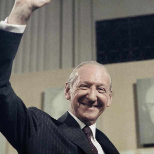 Ein lächelnder Mann im Anzug, Kurt Waldheim im Jahr 1986, hebt den Arm vor einem hellen Hintergrund.