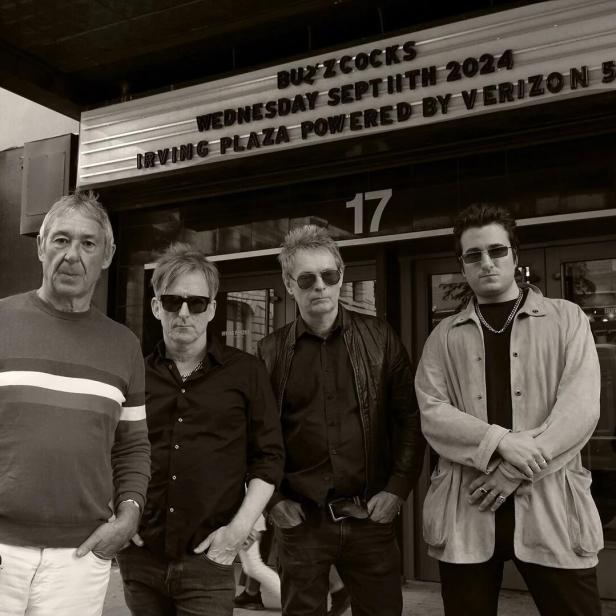 Die vierköpfige Band Buzzcocks in einem schwarzweißen Foto, hinten ein Lokal, das ihr Auftreten annonciert