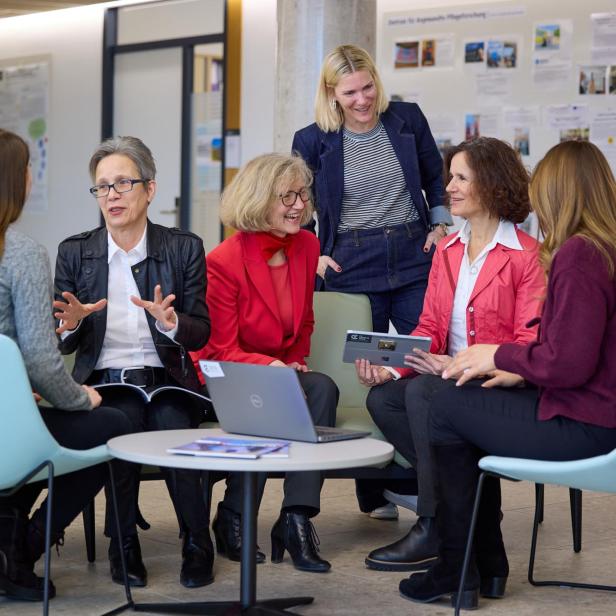 Sechs Frauen sitzen und stehen in einem Raum mit Informationsplakaten an der Wand, sie unterhalten sich in einer lockeren Runde um einen runden Tisch mit Laptop und Broschüren.