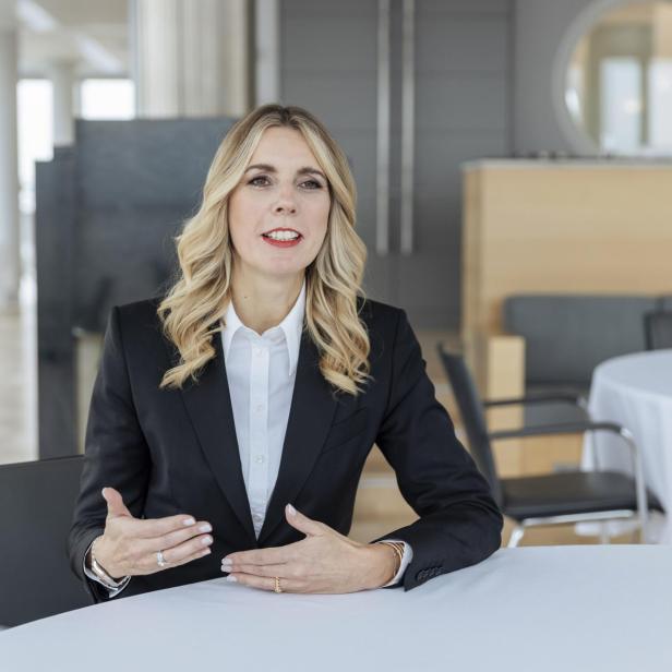 Frau mit langen blonden Haaren in schwarzem Blazer und weißem Hemd sitzt an einem runden Tisch in einem modernen Raum mit Spiegel und Stuhl im Hintergrund