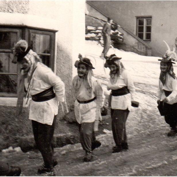 Mehrere Menschen in traditionellen Kostümen mit Masken und Hüten gehen in einer winterlichen Dorfszene hintereinander her.