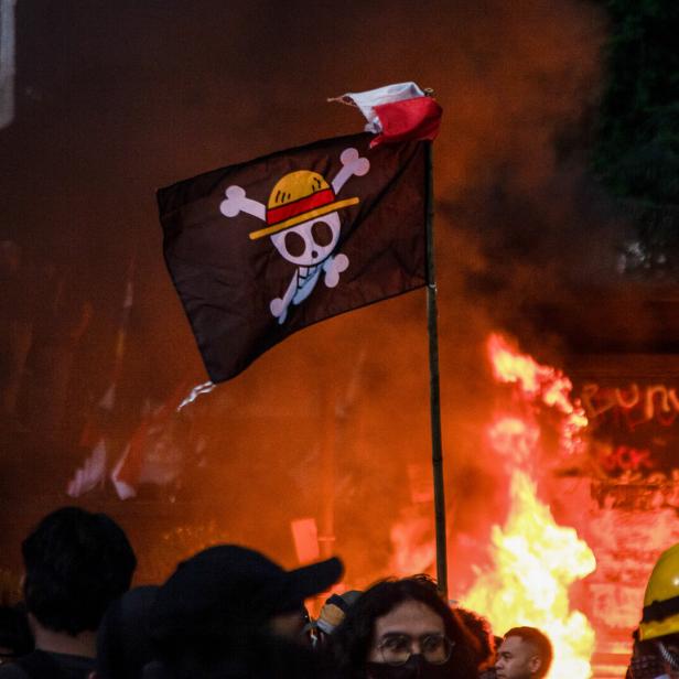 Menschenmenge vor einem großen Feuer, darüber eine schwarze Piratenflagge mit Strohhut und gekreuzten Knochen.
