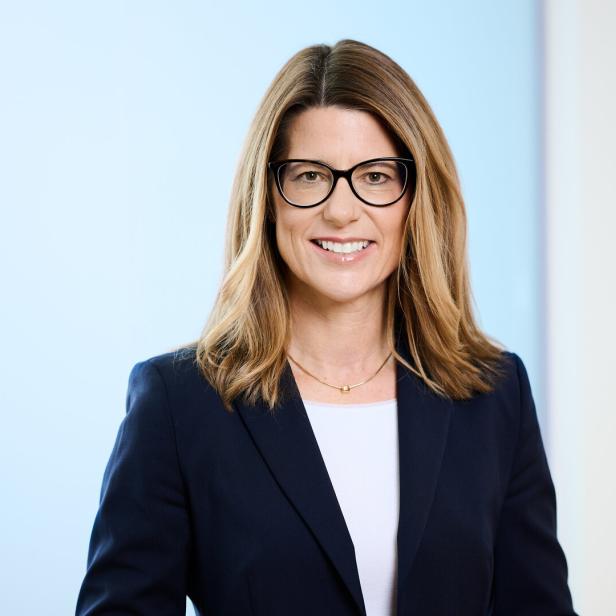 Eine Frau mit Brille, dunklem Blazer und blonden Haaren lächelt in die Kamera.