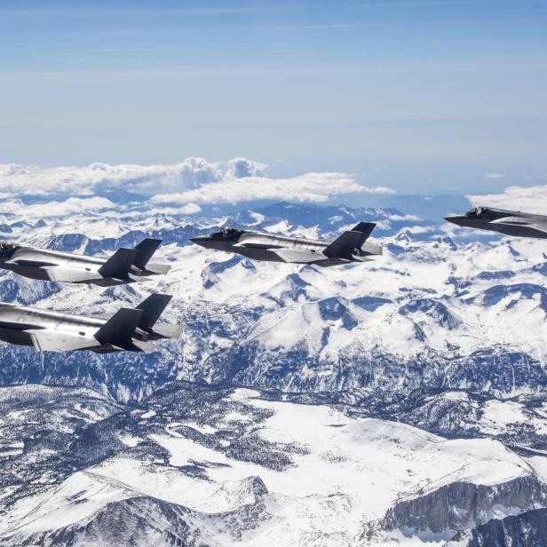 Vier moderne Kampfjets fliegen in Formation über schneebedeckten Bergen unter klarem Himmel