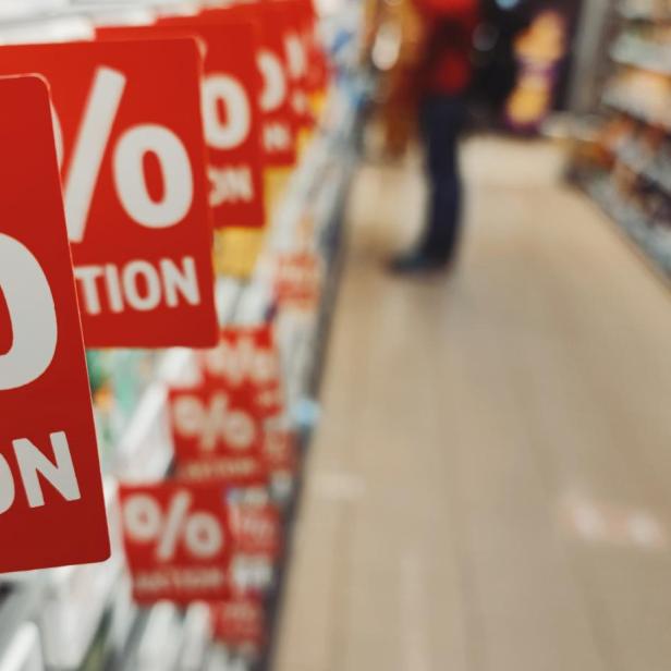 Rote Schilder mit Prozentzeichen und „Aktion“ hängen an einem Supermarktregal, im Hintergrund Regale und eine Person.