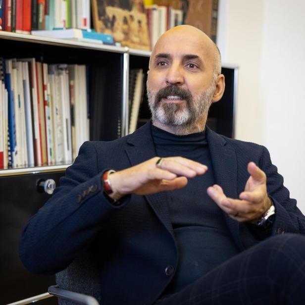 Ein Mann mit Bart und Glatze sitzt vor einem Bücherregal und gestikuliert mit den Händen, als ob er etwas erklärt.