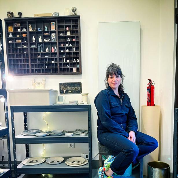 Eine Frau sitzt in einem Atelier auf einem Hocker, umgeben von Regalen mit Keramik und kleinen Figuren.