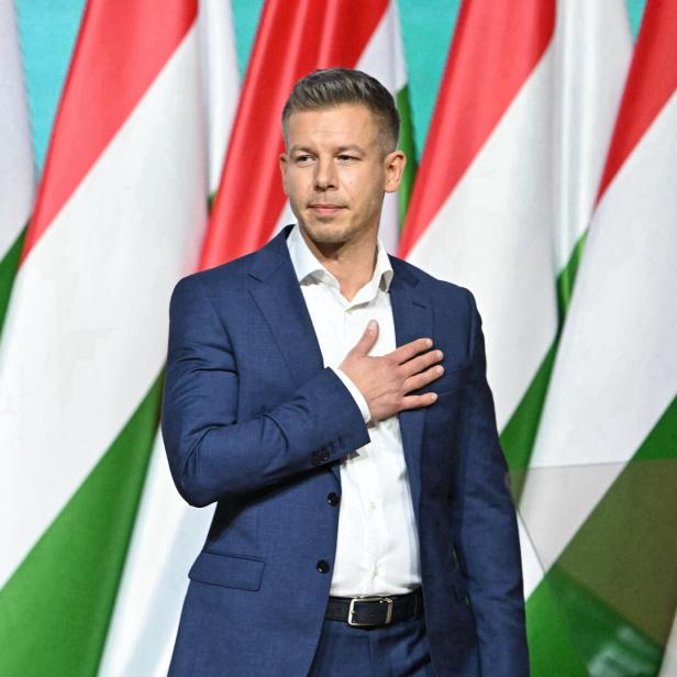 Peter Magyar legt seine Hand auf sein Herz, er steht vor Ungarn-Flaggen.