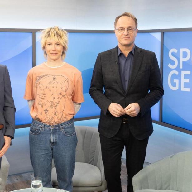 Drei Personen stehen vor einem blauen Hintergrund mit dem Schriftzug 'SPONTAN GEFRAGT', zwei tragen Anzüge, eine Person trägt ein T-Shirt und Jeans