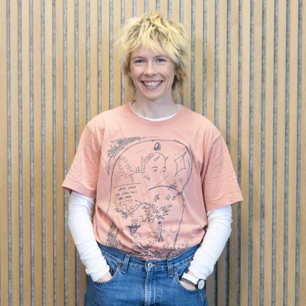 Person mit blonden, zerzausten Haaren trägt ein rosa T-Shirt mit schwarzem, skizzenhaftem Motiv über einem weißen Langarmshirt und blaue Jeans vor einer vertikal gestreiften Wand