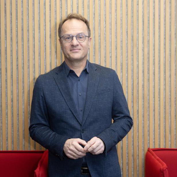 Mann in blauem Anzug und Hemd steht zwischen zwei roten Sesseln vor einer Wand mit vertikalen Holzleisten
