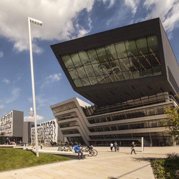 Modernes, kantiges Gebäude mit großer Glasfront und mehreren Stockwerken vor bewölktem Himmel und Personen auf dem Vorplatz
