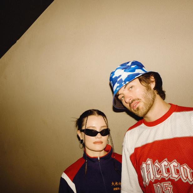Ein junges Pop-Duo, sie mit dunklen Sonnenbrillen, er mit blau-weißem Freizeithut, blickt in einem Raum mit schrägem Lichteinfall etwas misstrauisch in die Kamera.