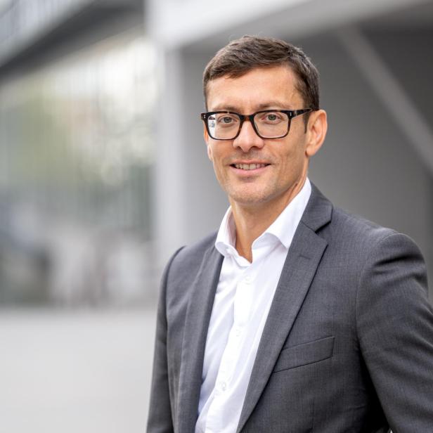 Mann mit dunklem Anzug und weißem Hemd steht vor unscharfem, modernem Gebäude
