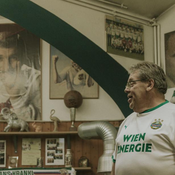 Fanclub-Präsident Martin Reiser in Rapid-Dress steht in seinem Vereinslokal, an der Wand etliche Poster und Fanschals