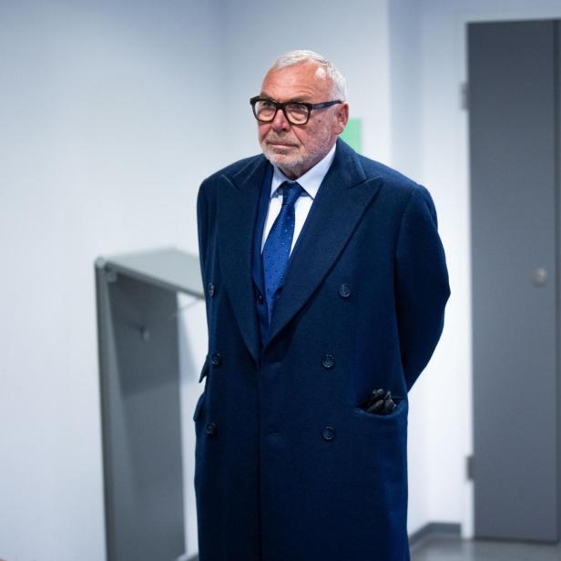 Älterer Mann mit Brille, dunklem Mantel und Krawatte vor dunkler Tür.