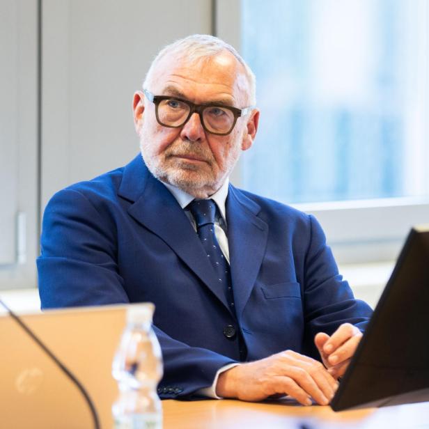 Ein älterer Mann im blauen Anzug sitzt an einem Tisch mit Laptop und Monitor.