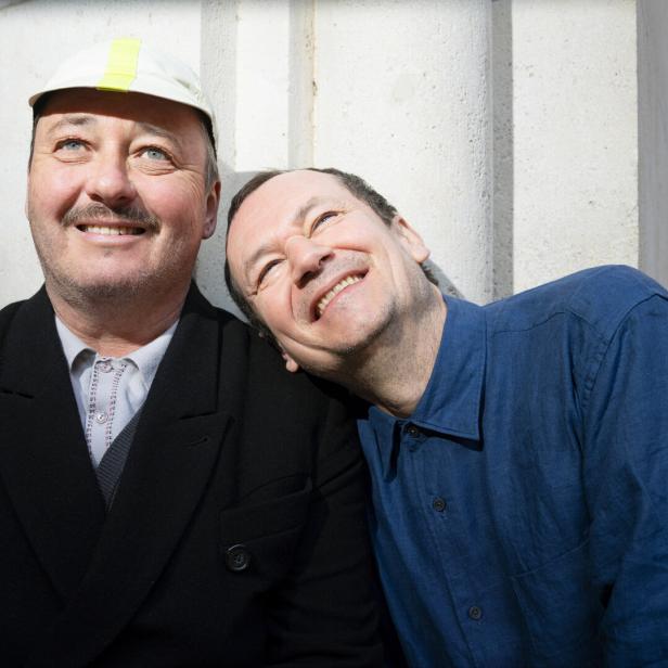 Die beiden Attwenger-Musiker Hans Peter Falkner und Markus Binder lehnen nebeneinander an einer Mauer und lächeln