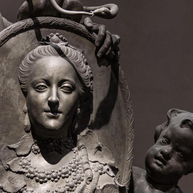Detail einer Skulptur mit einer Figur in aufwändiger Kleidung und Perlenketten vor einem Schild, darüber eine Schlange und eine Hand