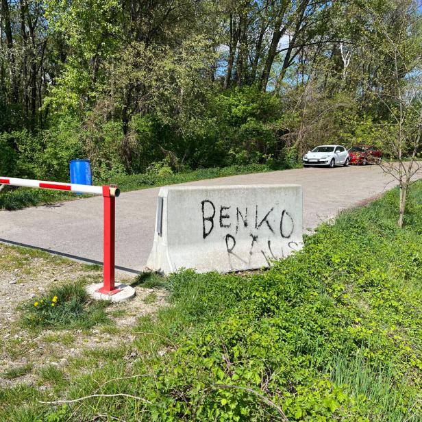 Eine rot-weiße Schranke und eine Betonbarriere mit Graffiti versperren eine Straße im Grünen, im Hintergrund stehen zwei Autos.