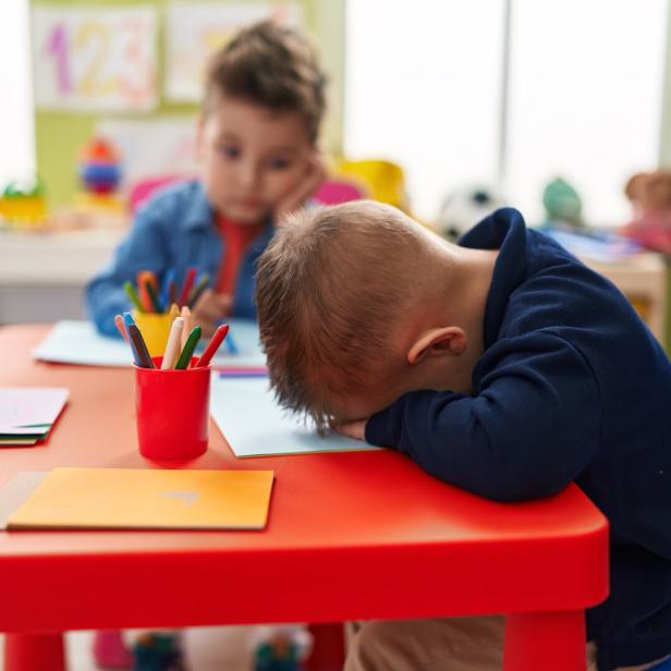 Zwei Kinder sitzen an einem Tisch, eines legt den Kopf erschöpft auf die Arme, das andere malt mit Buntstiften.