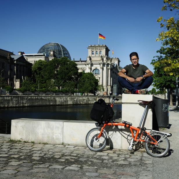 Der Autor Hasnain Kazim sitzt auf einem Mauervorsprung, im Hintergrund ist der Berliner Reichstag zu sehen, im Vordergrund steht ein Falt-Rad