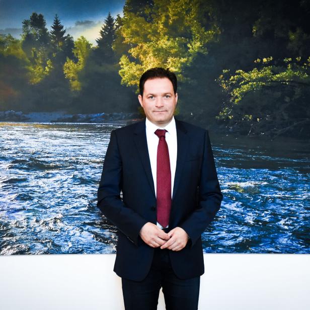Ein Mann im Anzug mit roter Krawatte, Klimaminister Totschnig, steht vor einem großen Wandbild mit Fluss und bewaldeter Landschaft.