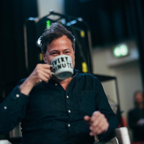 Regisseur Milo Rau trinkt mit erheiterter Miene aus einer beschrifteten Tasse in einem weitläufigen Innenraum.