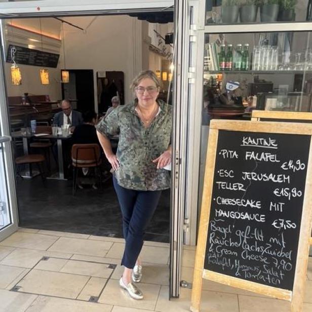 Frau steht am Eingang eines Cafés neben einer Tafel mit Speisekarte, Gäste sitzen im Hintergrund.
