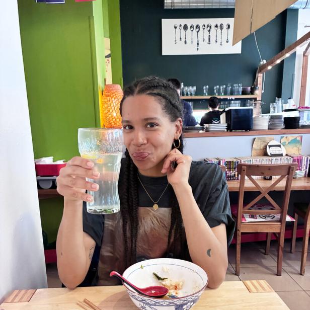 Rebecca Horner sitzt an einem Tisch, vor sich asiatische Speisen, in der Hand ein großes Glas Soda-Zitron