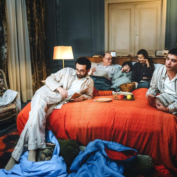 Ein Kind, zwei junge Männer und ein älteres Paar sitzen und liegen gemeinsam auf einem rot bezogenen Bett in einer Wohnung.