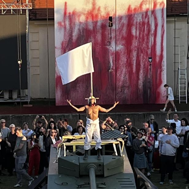 Halbnackte junge Schwertschluckerin steht mit weißer Fahne in einem Schlosshof auf einem Panzer, im Hintergrund ein monumentales rotgefrbtes Bild