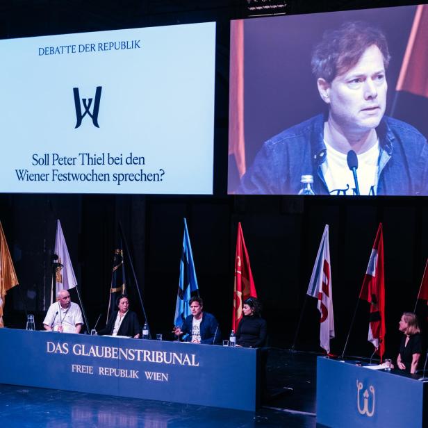 Foto eines beflaggten, mit fünf Menschen besetzten Podiums, auf einem Videoscreen dahinter sieht man Festwochenchef Milo Rau in Großaufnahme.