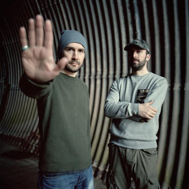 Zwei ernst dreinblickende Musiker in einem kargen Innenraum, der eine mit der verschränkten Armen, der andere hält die offene Hand in die Kamera, als wollte er das Foto untersagen. 