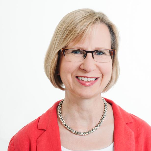 Oberkörper einer Person mit kurzen blonden Haaren, rotem Blazer, weißem Oberteil und silberner Kette vor weißem Hintergrund