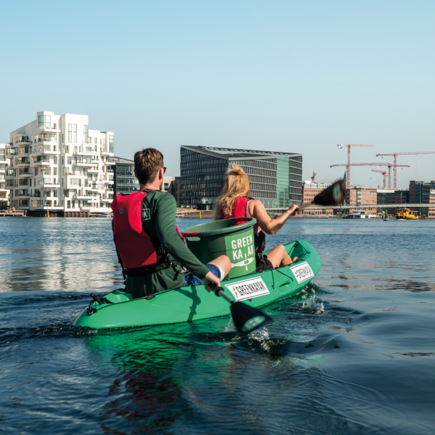 Zwei Personen in grünem Kajak auf ruhigem Wasser vor moderner Stadt mit mehreren Baukränen