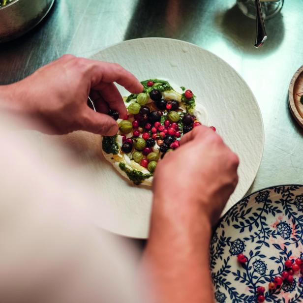 Hände dekorieren einen Teller mit einer Creme und verschiedenen Beeren und Kräutern auf einem Holztisch mit weiteren Tellern und Kräutern